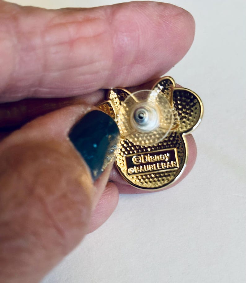 May include: Gold-colored Mickey Mouse pin with a clear plastic backing. The back of the pin has the text "&copy;Disney  &copy;AUBLEEBAR".