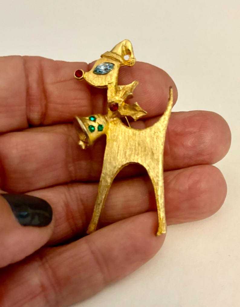 May include: Gold-tone reindeer brooch with red, blue, and green rhinestones. The reindeer is wearing a Santa hat and has a bell around its neck.