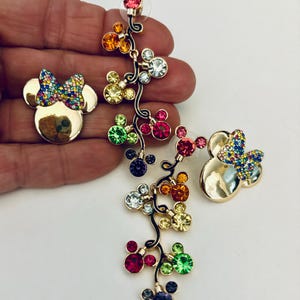 May include: A pair of gold-tone dangle earrings featuring a cluster of colorful crystal Mickey Mouse heads. The earrings are designed with a dangling chain of Mickey Mouse heads with different colored crystals. The earrings are perfect for adding a touch of Disney magic to any outfit.