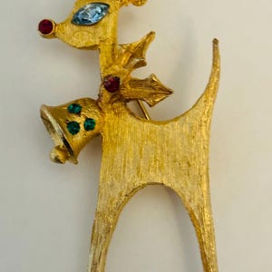 May include: A gold-toned reindeer brooch with red and green rhinestones. The reindeer is wearing a Santa hat and has a bell around its neck.