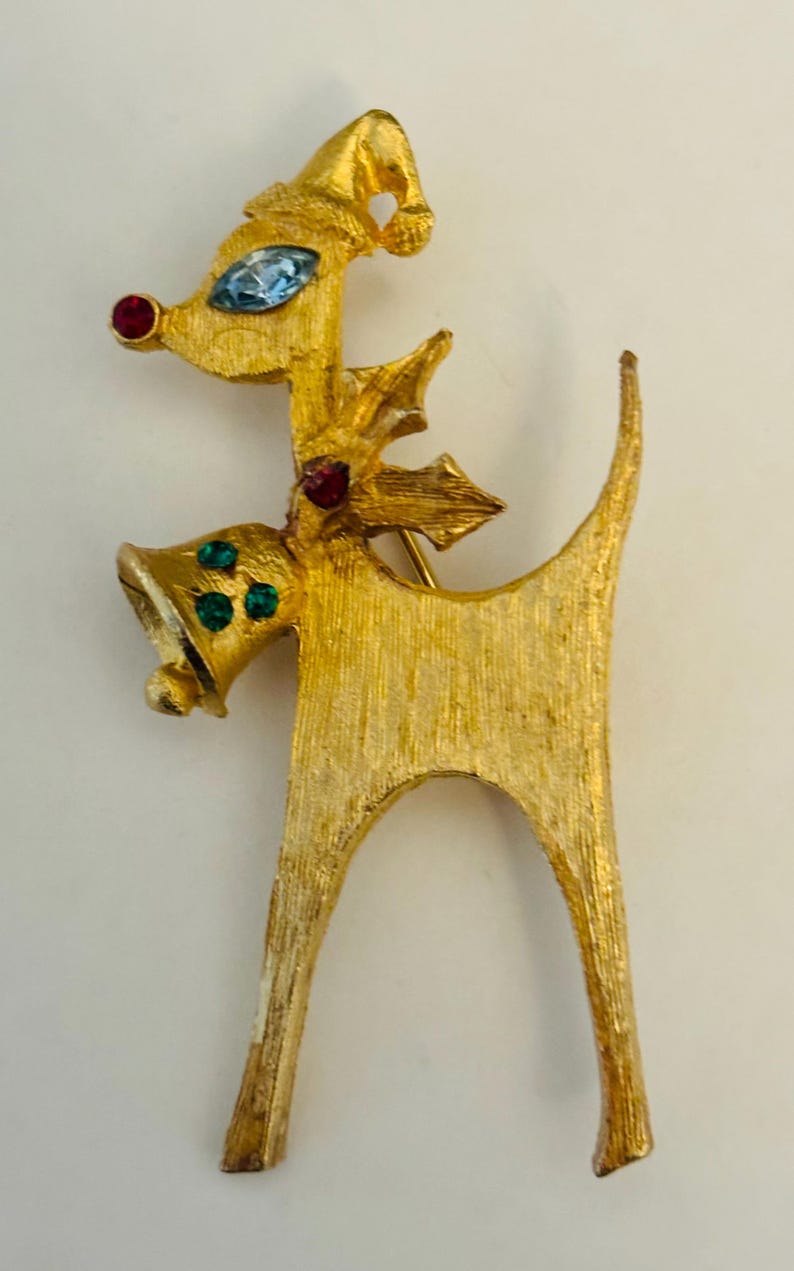 May include: A gold-toned reindeer brooch with red and green rhinestones. The reindeer is wearing a Santa hat and has a bell around its neck.