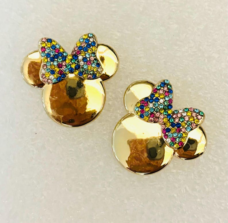 May include: A pair of gold-tone earrings with a Minnie Mouse design. The earrings feature a bow with colorful rhinestones.