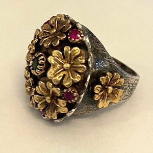 May include: A silver ring with a textured band and a large, round, gold-toned top. The top is decorated with several small, gold-toned flowers and red and green gemstones.