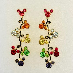 May include: A pair of dangle earrings with a Mickey Mouse head design. The earrings are made of gold-tone metal and feature a cascade of colorful crystals in a rainbow pattern.