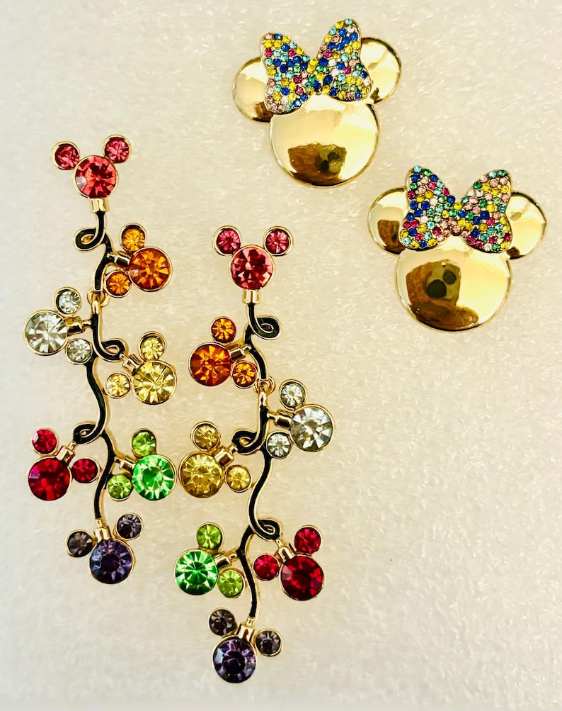 May include: A pair of gold-tone dangle earrings with a black stem and colorful crystal Mickey Mouse heads. The earrings are accompanied by two gold-tone Mickey Mouse head pins with colorful crystal bows.