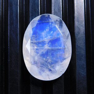 Puede incluir: Una piedra lunar ovalada con una superficie facetada. La piedra muestra un cautivador juego de colores, con un centro azul brillante y un exterior blanco lechoso translúcido. La gema se encuentra sobre un fondo oscuro y texturizado.