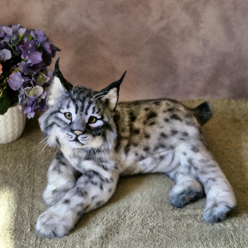 May include: A detailed plush lynx toy with a spotted grey and white coat, realistic facial features, and pointed ears. The toy cat is lying down on a neutral surface, with a blurred floral arrangement in the background.