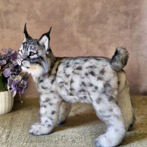May include: A plush lynx figurine with a spotted grey and white coat, upright ears, and a short tail. The handmade animal stands on four legs, with a detailed face and whiskers. The figurine is a decorative item.