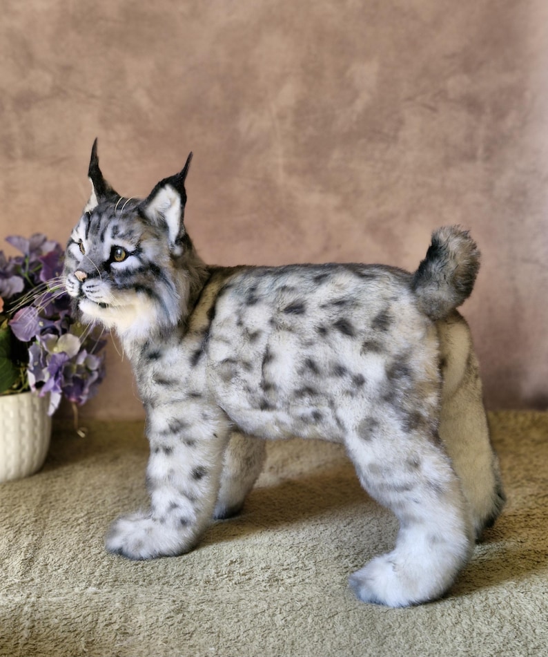 May include: A plush lynx figurine with a spotted grey and white coat, upright ears, and a short tail. The handmade animal stands on four legs, with a detailed face and whiskers. The figurine is a decorative item.