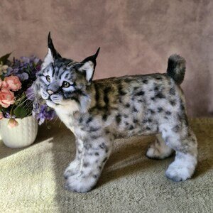 May include: A detailed plush lynx toy, featuring a spotted grey and tan coat, black-tipped ears, and yellow eyes. The handmade animal stands on four paws, with a fluffy tail. The soft toy is a realistic representation of a wild cat, suitable for display.
