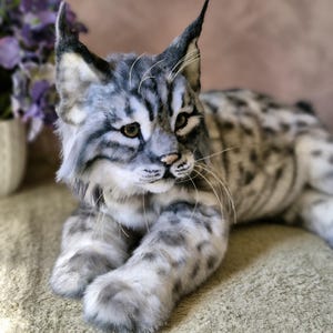 May include: A detailed plush lynx cat with grey and white fur, featuring dark stripes and spots. The cat has pointed ears, a pink nose, and whiskers. It is lying down on a textured surface, displaying its realistic features.