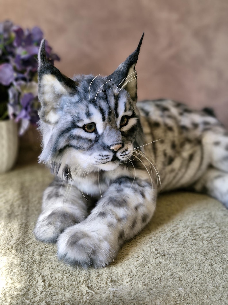 May include: A detailed plush lynx cat with grey and white fur, featuring dark stripes and spots. The cat has pointed ears, a pink nose, and whiskers. It is lying down on a textured surface, displaying its realistic features.