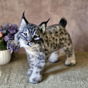 May include: A handcrafted bobcat sculpture with a realistic appearance. The bobcat features a grey and white coat with black markings, erect ears, and a small tail. It stands on a light green surface.