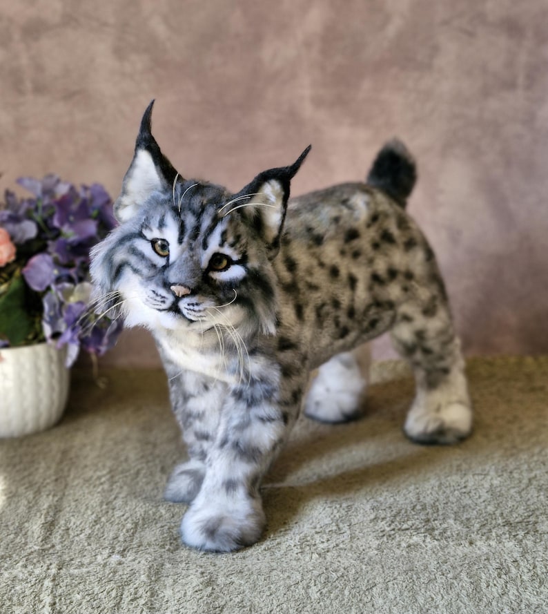 May include: A handcrafted bobcat sculpture with a realistic appearance. The bobcat features a grey and white coat with black markings, erect ears, and a small tail. It stands on a light green surface.