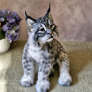 May include: A detailed, plush bobcat figure with a grey and white spotted coat, upright ears, and realistic facial features. The handmade cat is posed sitting, displaying its fluffy paws and textured fur. A decorative item, suitable for display.