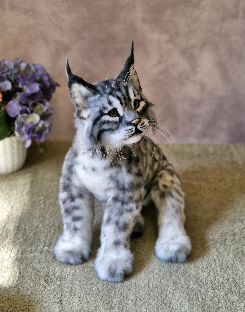 May include: A detailed, plush bobcat figure with a grey and white spotted coat, upright ears, and realistic facial features. The handmade cat is posed sitting, displaying its fluffy paws and textured fur. A decorative item, suitable for display.
