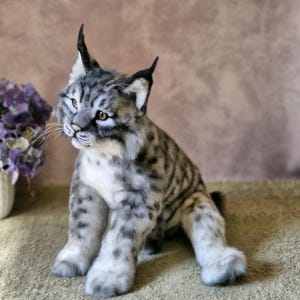 May include: A detailed, plush bobcat toy with a grey and white striped coat, black ear tufts, and yellow eyes. The handmade cat is posed sitting upright, displaying its realistic fur and features. A decorative item for cat enthusiasts.