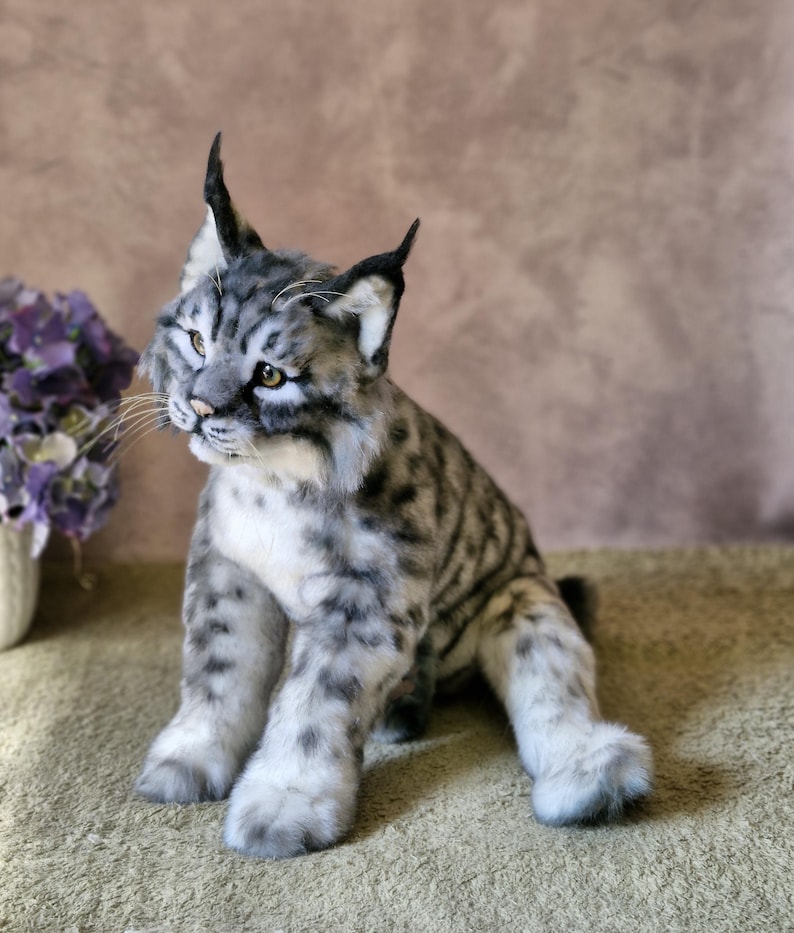 May include: A detailed, plush bobcat toy with a grey and white striped coat, black ear tufts, and yellow eyes. The handmade cat is posed sitting upright, displaying its realistic fur and features. A decorative item for cat enthusiasts.