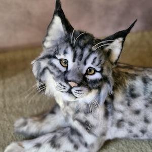May include: A detailed plush lynx toy with grey and white fur, featuring black markings and distinctive tufted ears. The cat has yellow eyes and a pink nose, resting on a neutral surface. The toy is designed to resemble a real lynx.