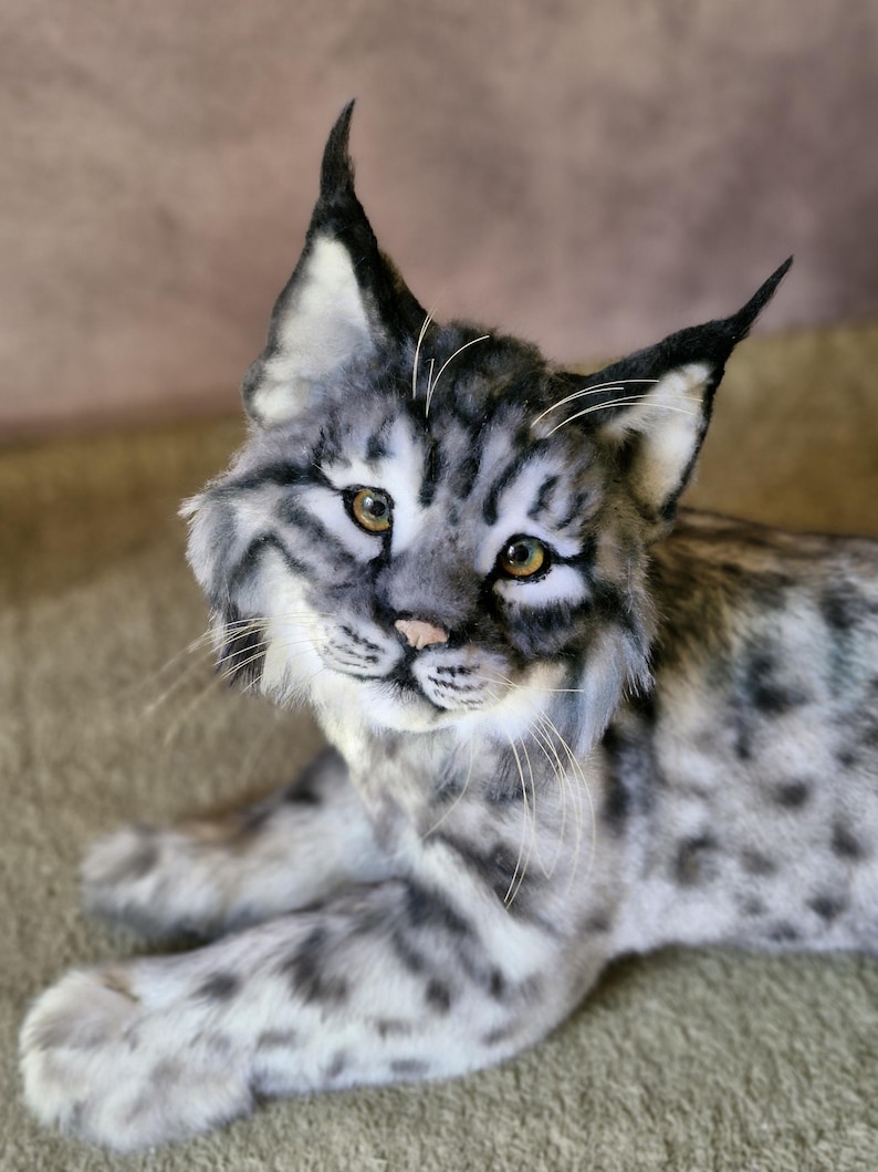 May include: A detailed plush lynx toy with grey and white fur, featuring black markings and distinctive tufted ears. The cat has yellow eyes and a pink nose, resting on a neutral surface. The toy is designed to resemble a real lynx.