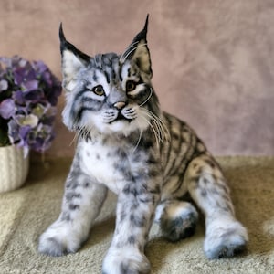 May include: A lifelike plush bobcat toy. It has a grey and white striped coat, distinctive pointed ears, and a focused expression. The bobcat is seated, displaying its paws and textured fur. The toy is designed to resemble a real bobcat.