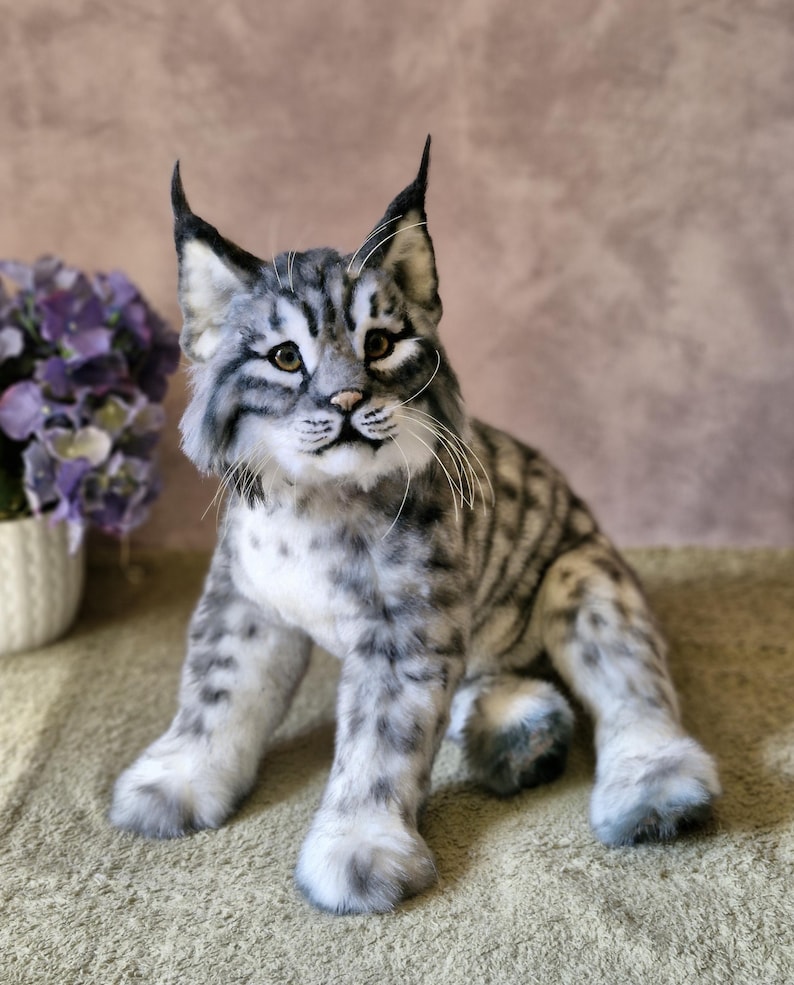 May include: A lifelike plush bobcat toy. It has a grey and white striped coat, distinctive pointed ears, and a focused expression. The bobcat is seated, displaying its paws and textured fur. The toy is designed to resemble a real bobcat.