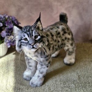 May include: A detailed plush lynx toy, featuring a spotted grey and white coat, erect ears, and a short tail. The handcrafted animal has realistic facial features, including whiskers and brown eyes, and stands on four paws.
