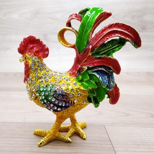 Könnte beinhalten: Eine farbenfrohe Metallfigur eines Hahns mit gelbem Körper, roten und grünen Schwanzfedern und einem Juwelendesign. Der Hahn steht auf einer Holzoberfläche.