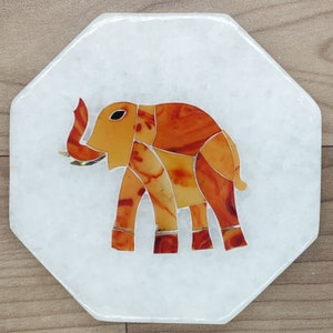 May include: White octagonal coaster with an inlaid elephant design. The elephant is composed of orange and yellow pieces with silver accents. The coaster is made of a white stone material and is set against a wooden surface.