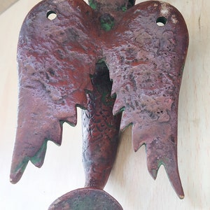 May include: A vintage, bronze-colored metal wall hanging in the shape of a stylized bird or angel. The piece features textured wings, a central body, and a rounded base with two holes. The metal has a weathered appearance with hints of green.