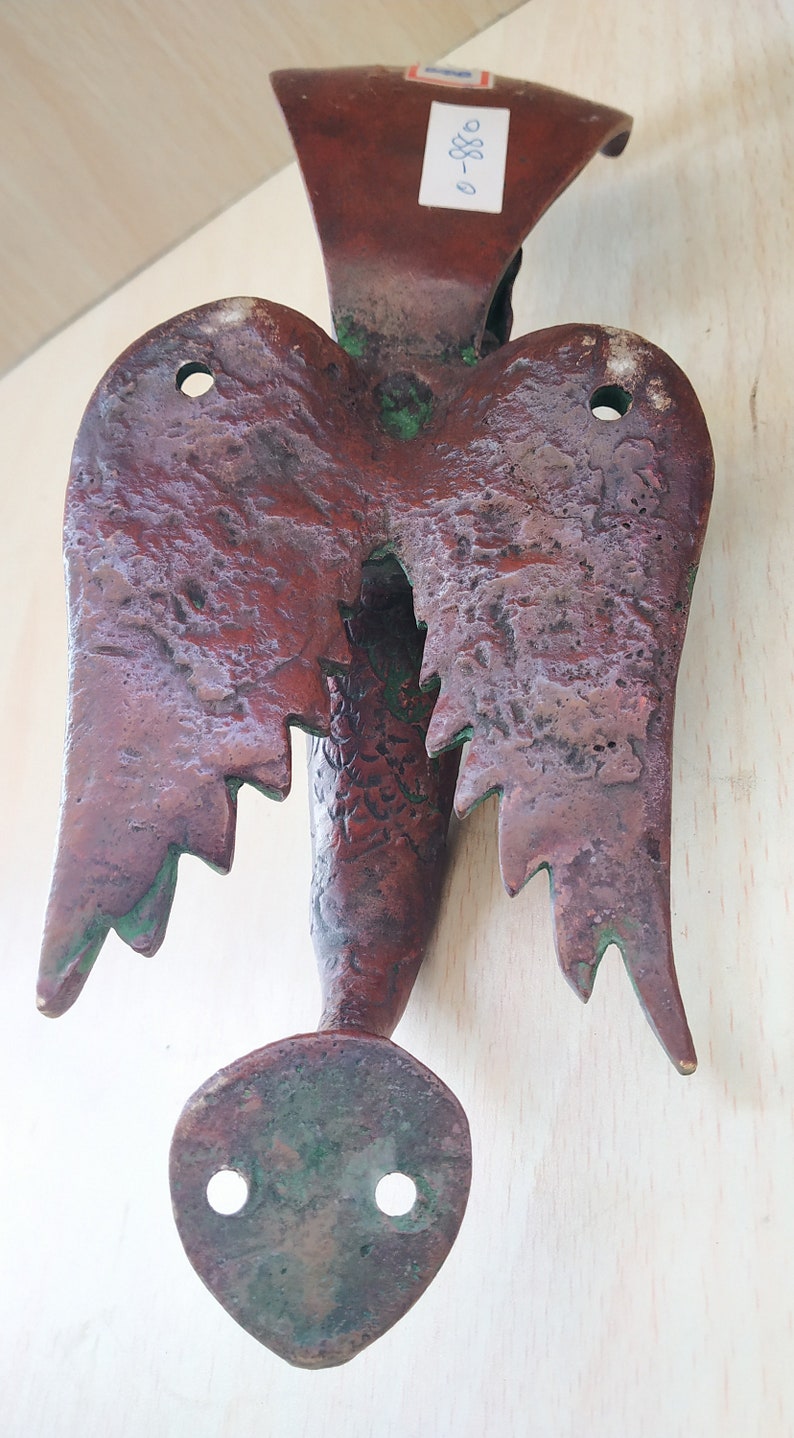 May include: A vintage, bronze-colored metal wall hanging in the shape of a stylized bird or angel. The piece features textured wings, a central body, and a rounded base with two holes. The metal has a weathered appearance with hints of green.