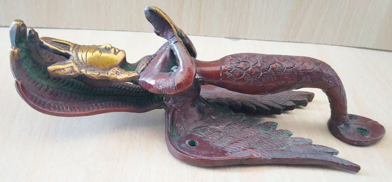 May include: A bronze-colored door handle featuring a mermaid design. The handle has a detailed mermaid figure with a golden face and flowing hair, wings, and a scaled tail. The handle is likely made of metal and has a vintage or antique aesthetic.