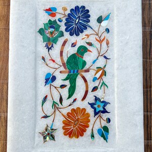 Puede incluir: Una bandeja de mármol blanco con un diseño floral incrustado con piedras de colores. El diseño presenta un pájaro posado en una rama con flores y hojas.