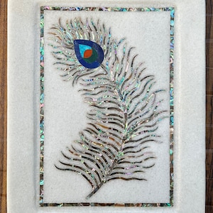 Puede incluir: Bandeja decorativa de mármol blanco con diseño de pluma de pavo real. La pluma está incrustada con piezas de concha iridiscente y presenta un ojo azul, naranja y verde. La bandeja tiene forma rectangular con esquinas redondeadas y borde de mosaico.