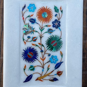 Puede incluir: Una bandeja de mármol blanco con un diseño floral incrustado con piedras de colores. El diseño presenta flores azules, verdes, naranjas y blancas con detalles intrincados.