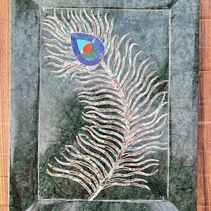 Puede incluir: Una bandeja de mármol verde con una incrustación de pluma de pavo real hecha de nácar iridiscente. La pluma está detallada con un ojo azul, rojo y verde.