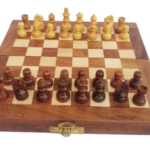 May include: A wooden chess set with a folding board. The board is closed and the pieces are arranged in their starting positions. The pieces are made of wood and are painted in traditional colors. The board is made of dark wood with light squares and light wood with dark squares.