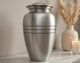 May include: A brushed silver metal cremation urn with a smooth, rounded body and a matching lid. The urn features three horizontal engraved lines encircling the center of the vessel, providing a subtle decorative detail to the metallic finish.