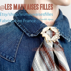 Peut inclure: Un foulard vintage avec un fermoir décoratif, présentant un motif géométrique marron, gris et blanc. Le foulard est associé à une chemise en jean. Le texte "@LES MAUVAISES FILLES" et "Fabriqué en France" sont visibles.