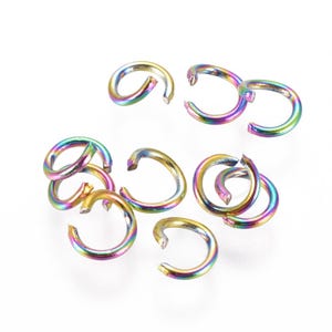Könnte beinhalten: Eine Sammlung schillernder Sprungringe in verschiedenen Größen. Die offenen Ringe zeigen ein Farbspektrum, darunter Gold, Lila, Blau und Grün, wodurch ein Regenbogeneffekt entsteht. Die Schmuckzubehörteile sind auf einer weißen Oberfläche verstreut.