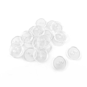 May include: A pile of clear, transparent buttons, each with two holes. The circular buttons are made of a clear material, possibly plastic. They are scattered on a white background, suitable for sewing or craft projects.