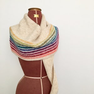 Può includere: Una sciarpa di maglia color crema con un motivo a righe arcobaleno. La sciarpa è drappeggiata su un manichino.