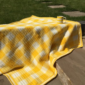 May include: A yellow and white gingham crochet throw blanket with a white border. The blanket is folded and draped over a set of stone steps. A yellow and white striped mug is sitting on top of the blanket.