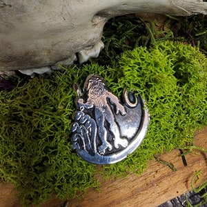 May include: A silver pendant featuring a mythological figure with a lion's head and a human body, holding a sword. The pendant is set against a backdrop of green moss, wood, and a bone.