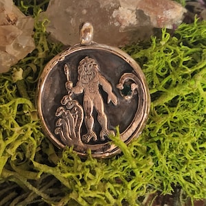 May include: A round, silver-toned pendant featuring a detailed relief of a mythical figure with a lion's mane, holding a spear, and standing above serpent-like creatures. The pendant has a dark background and a small green gem. The pendant is displayed on a bed of green moss.