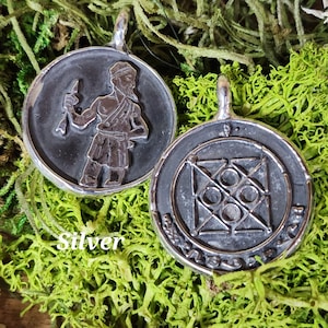 May include: Two silver pendants, one with a detailed image of a person holding a weapon and the other with a geometric design and text.