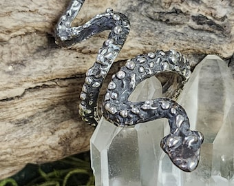 May include: A silver-toned snake ring with a textured, scale-like design. The ring is coiled and features a detailed snake head. The ring is displayed on a clear crystal, with a wooden background.