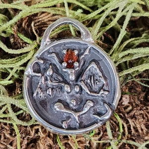 May include: A detailed, oxidized silver pendant with a loop at the top. The pendant features an intricate design with a central, teardrop-shaped, red gemstone. The design includes raised, organic shapes and textures, creating a unique, artistic piece.
