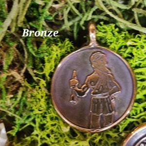 May include: A bronze pendant featuring a detailed image of a person holding a staff. The pendant is oval shaped and has a smooth, polished finish.
