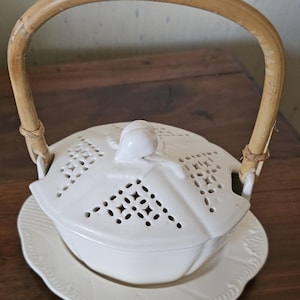 Può includere: Un piatto da portata in ceramica bianca con coperchio traforato e manico in bambù. Il piatto poggia su un piattino abbinato con bordo smerlato. Il manico è fissato con spago intrecciato.